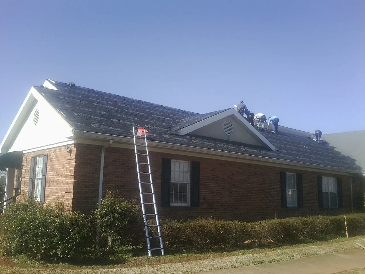 Crew tearing shingles off a brick ranch roof with a ladder set — Metal Roof Repair work by our crew in Lake Toxaway