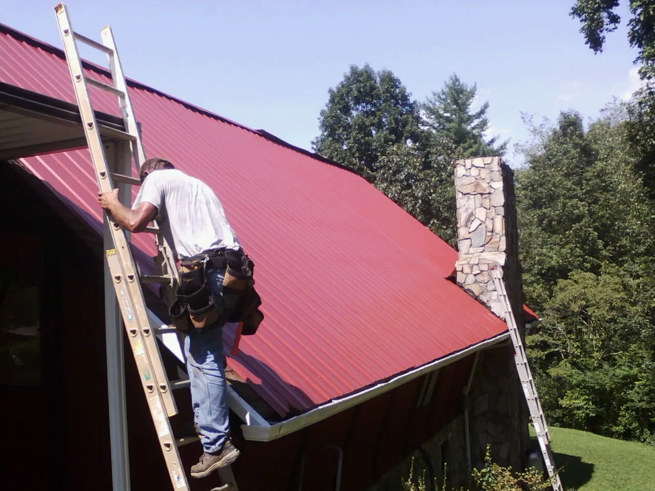 Roofer on a ladder beside a red metal roof and stone chimney — Soffit & Fascia Repair by Roofing Brothers in Lake Toxaway